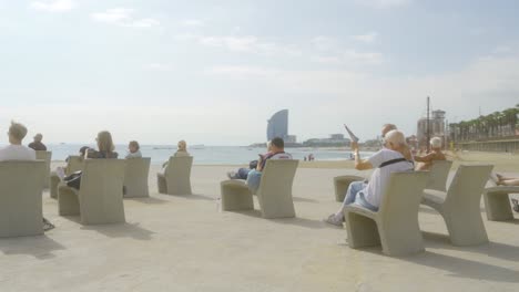 People-sitting-on-stone-sunbeds-at-Barceloneta-Beach-in-Barcelona,-Spain,-on-sunny-day-at-the-ocean