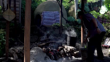 Man-at-village-hut-boiling-water-in-metal-buckets-on-a-smoldering-bonfire,-Medium-shot