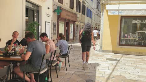 Following-a-tourist-walking-a-white-stoney-streets-in-Zadar,-among-people-sitting-and-eating-on-terrace-of-a-restaurants-and-street-turning-past-shops-and-pharmacy-on-a-sunny-summer-day