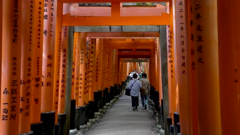 Touristen-Gehen-Durch-Torii-Tore-Mit-Kanji-Schrift-Darauf-Bei-Fushimi-Inari-Taisha