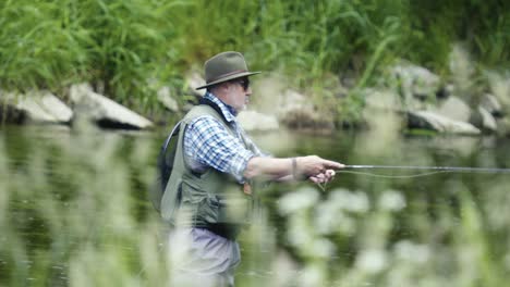 Man-fly-fishing-in-a-river,-wearing-a-hat-and-sunglasses,-casting-bait,-close-up-shot-with-grass-in-the-foreground