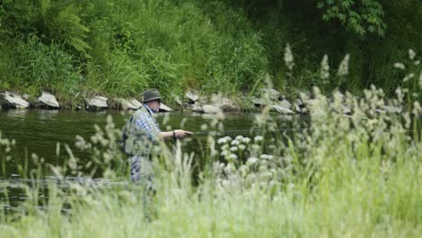A-man-wearing-a-hat-and-sunglasses-stands-in-a-river,-fly-fishing