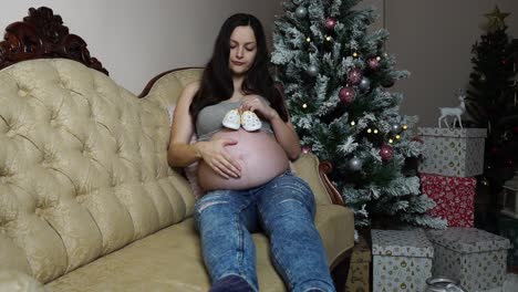 Pregnant-caucasian-woman-hold-baby-shoes-and-sit-on-couch-near-Christmas-tree