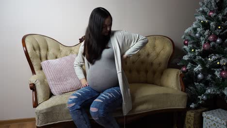 Pregnant-brunette-woman-sit-on-couch-during-Christmas-with-lower-back-pain