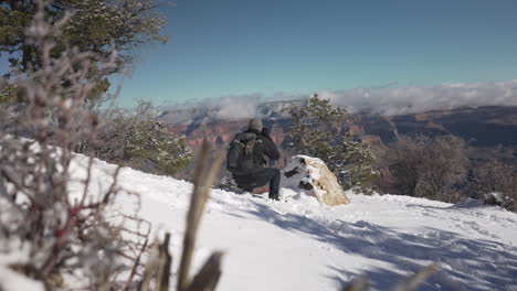 Man-crouching-down-in-the-snow-taking-a-photo-of-the-grand-canyon---winter,-arizona,-grand-canyon,-tourist,-photographer