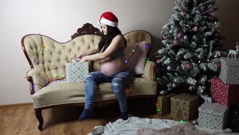 Pregnant-woman-with-Santa-hat-open-Christmas-gift-near-twinkle-lights-and-tree
