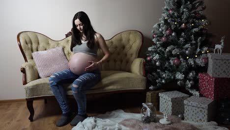 Pregnant-woman-sit-near-Christmas-tree-and-softly-touch-big-round-belly