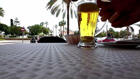 Close-up-of-a-hand-picking-up-a-beer-glass-on-a-table-with-a-palm-lined-street-in-the-background