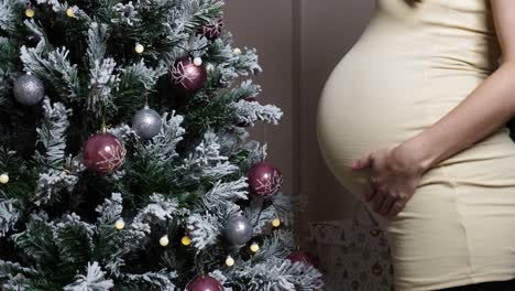Pregnant-woman-with-big-round-belly-stand-near-decorated-Christmas-tree