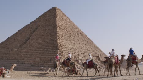 Turistas-Montando-Camellos-Recorren-Las-Pirámides-Egipcias,-Recorrido-Histórico-Y-Cultural,-Pirámides-De-La-Unesco-El-Cairo-Egipto