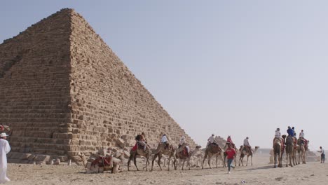 Turistas-Caminando-Alrededor-De-Las-Pirámides-Con-Camellos,-Un-Día-Soleado-En-Las-Pirámides-Egipcias,-Estructuras-Mundialmente-Famosas-El-Cairo,-Egipto