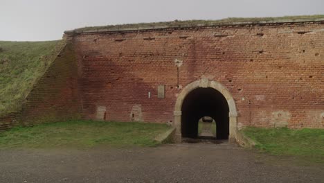 Close-up-of-the-old-brick-wall-of-the-Terezín-Fortress-with-a-small-arched-entrance,-surrounded-by-grass-and-a-clear-sky-above