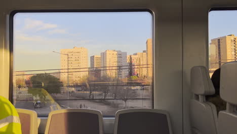 Through-train-window,-towering-city-houses-illuminated-by-evening-sun,-set-against-canvas-of-clear-sky