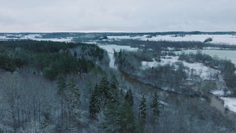 Aerial-establishing-view-of-valley-of-the-Abava-river-on-overcast-winter-day,-fields-covered-in-snow,-Abava-river-filled-with-dark-flood-water,-wide-drone-shot-moving-forward