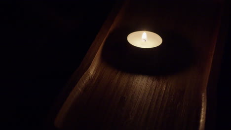 Single-tea-candle-burns-in-rich-brown-wooden-tray-in-dark-black-room