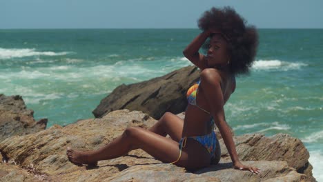Enjoying-the-sunshine,-an-African-girl-in-a-bikini-sits-atop-a-cliff,-her-natural-hair-framing-her-as-she-gazes-over-the-expanse-of-the-ocean