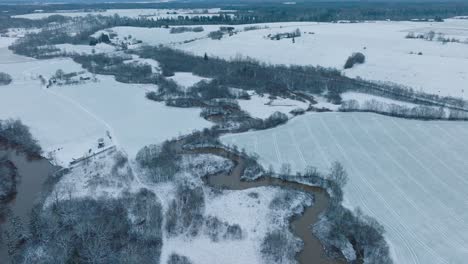 Aerial-establishing-view-of-valley-of-the-Abava-river-on-overcast-winter-day,-fields-covered-in-snow,-Abava-river-filled-with-dark-flood-water,-wide-drone-shot-moving-forward,-tilt-down