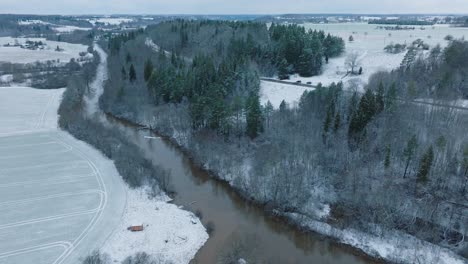 Aerial-establishing-view-of-valley-of-the-Abava-river-on-overcast-winter-day,-fields-covered-in-snow,-Abava-river-filled-with-dark-flood-water,-wide-drone-shot-moving-forward