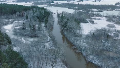 Aerial-establishing-view-of-valley-of-the-Abava-river-on-overcast-winter-day,-fields-covered-in-snow,-Abava-river-filled-with-dark-flood-water,-wide-drone-shot-moving-forward,-tilt-down