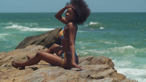 On-a-sunny-day,-an-African-girl-sits-on-a-cliff-overlooking-the-ocean,-wearing-a-bikini-and-letting-her-natural-hair-sway-in-the-breeze