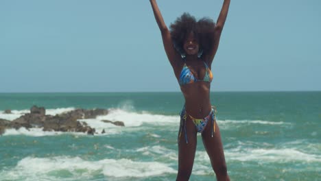 An-Einem-Hellen-Und-Sonnigen-Tag-Steht-Ein-Afrikanisches-Mädchen-Im-Bikini-Auf-Einer-Klippe,-Ihr-Natürliches-Haar-Tanzt-Im-Wind,-Während-Sie-Den-Ozean-Unter-Sich-Betrachtet