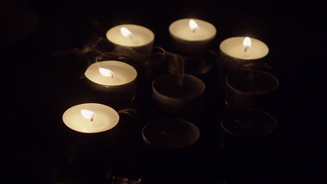Group-of-burning-tea-candles-on-reflective-tray-blown-out-to-darkness