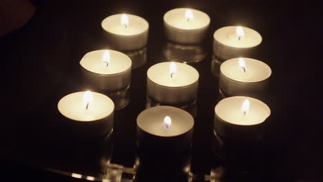 Nine-tea-candles-burn-flames-on-reflective-surface-in-dark-black-room