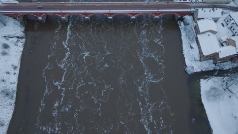 Aerial-establishing-view-of-Venta-river-rapids-during-winter-flood,-old-red-brick-bridge,-Kuldiga,-Latvia,-overcast-winter-day,-wide-birdseye-drone-shot-moving-forward