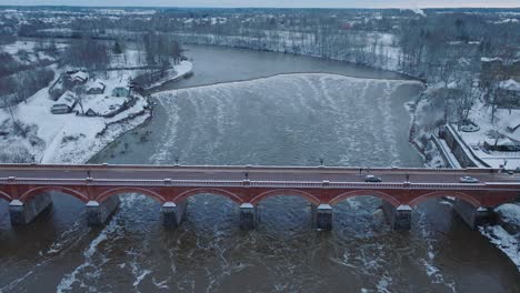 Aerial-establishing-view-of-Venta-river-rapids-during-winter-flood,-old-red-brick-bridge,-Kuldiga,-Latvia,-overcast-winter-day,-wide-birdseye-drone-shot-moving-forward