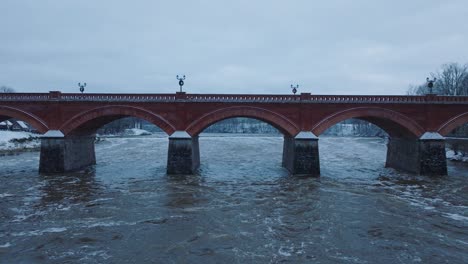Aerial-establishing-view-of-Venta-river-rapids-during-winter-flood,-old-red-brick-bridge,-Kuldiga,-Latvia,-overcast-winter-day,-wide-ascending-drone-shot-moving-forward