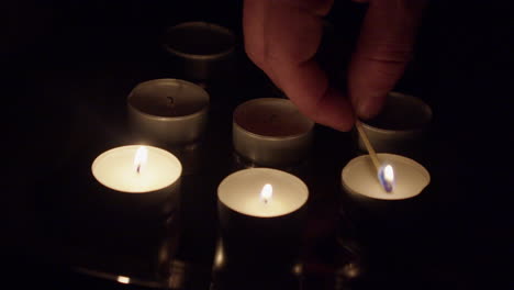 Fingers-hold-burning-wooden-match-to-light-tea-candles-in-dark-room