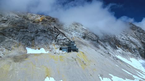 Vista-Aérea-De-La-Montaña-Zugspitze,-El-Pico-Más-Alto-De-Alemania,-Con-Nieve-Esporádica,-Cielo-Azul-Y-Nubes-En-El-Fondo