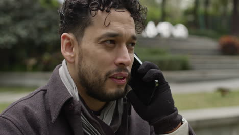 Close-up-slow-motion-of-caucasian-male-talking-at-phone-wearing-gloves-during-winter-season-sitting-outdoors-in-a-park-with-tree-in-residential-district