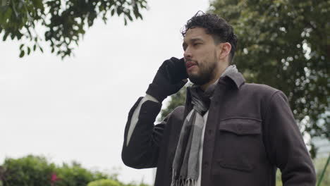 Caucasian-male-model-talking-at-phone-having-a-business-call-outdoors-with-tree-in-background-during-winter-season-wearing-gloves-and-scarf-modern-clothing-outfits