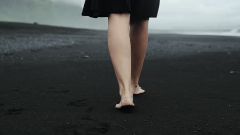 Füße-Gehen-Am-Schwarzen-Sandstrand,-Island,-Schöne-Frau-Im-Schwarzen-Kleid,-Zeitlupenverfolgung,-Dramatische-Meereslandschaft