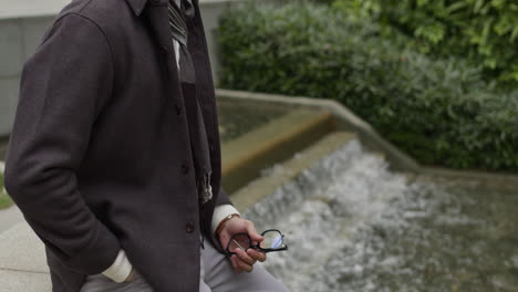 Caucasian-modern-male-with-trendy-clothes,-man-model-sitting-in-front-of-a-water-river-holding-his-glasses-in-his-hands-while-thinking