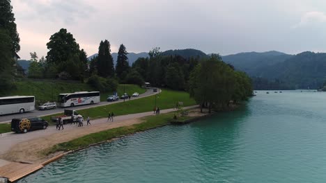 Drone-backwards-ascending-flying-view-of-lake-Bled-and-tourists-coming-from-a-bus