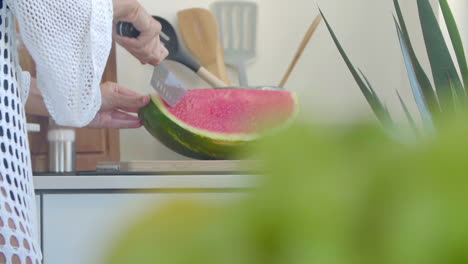 Close-up-of-a-woman-in-a-white-dress-slices-a-ripe-watermelon-on-a-wooden-board-with-a-large-knife-in-the-kitchen