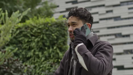 Caucasian-male-model-wearing-gloves-scarf-and-winter-clothing-in-a-phone-call-with-modern-smartphone-with-concrete-city-building-in-background,-slow-moition