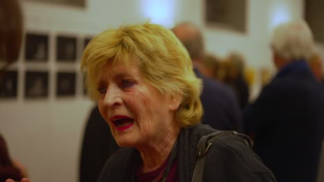 White-senior-woman-with-painted-blond-hair-talking-during-an-art-event-opening