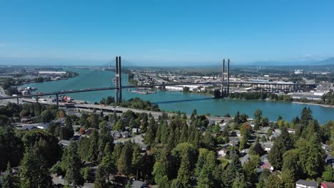 Drone-shot-showing-Alex-Fraser-Bridge-with-blue-sky,-trees,-residential-homes-and-river