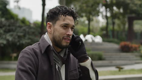 Push-back-shot-of-handsome-Hispanic-ethnicity-man-talks-positively,-confident-on-the-phone-outdoors-in-the-park
