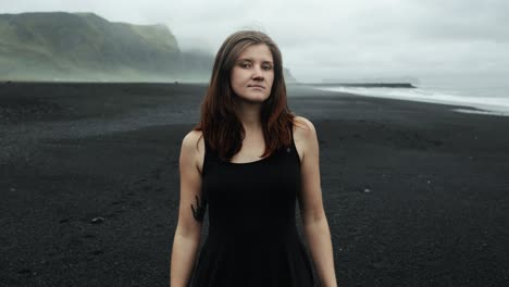 Young-beautiful-woman-in-black-dress-at-black-sand-beach-Iceland,-looking-into-camera-model-posing,-slow-motion,-dramatic-waves-seascape