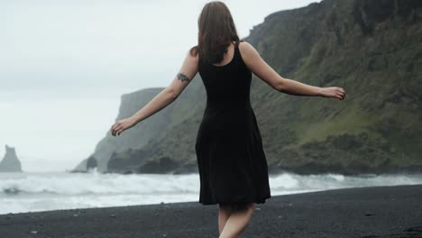 Young-beautiful-woman-in-black-dress-dancing-spinning-on-black-sand-beach-Iceland,-slow-motion,-dramatic-waves-seascape