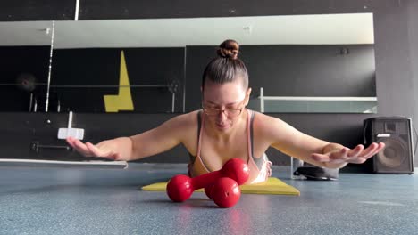 A-woman-lying-face-down-on-the-mat-in-the-gym,-exercising-with-her-hands-in-the-air,-a-woman-with-glasses-doing-sports