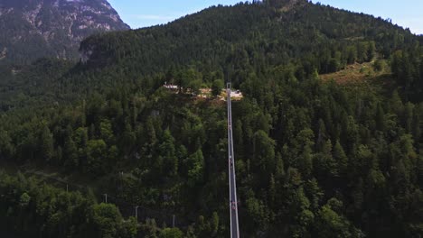 Panoramic-aerial-view-of-the-world's-longest-pedestrian-suspension-bridge-leading-to-Ehrenberg-Castle-in-Austria,-over-a-busy-road,-surrounded-by-a-forest-of-trees