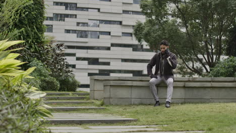 Caucasian-male-well-dressed-talking-at-phone-sitting-in-a-bench-of-a-residential-area-of-smart-city-with-buildings-on-background-in-green-park-area-with-tree-and-vegetation