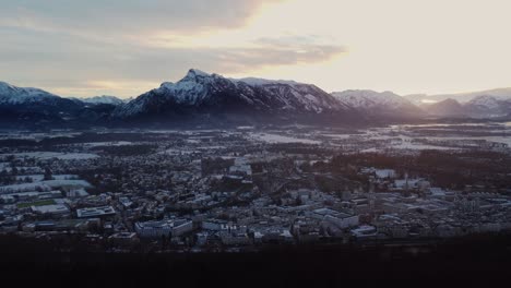 Snow-covered-Salzburg-cityscape,-castle-and-Mountain-alps-at-sunset,-aerial