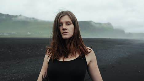 Young-beautiful-woman-in-black-dress-dancing-on-black-sand-beach-Iceland,-slow-motion-looking-into-camera,-dramatic-waves-seascape