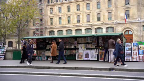 Parisian-kiosk-with-paintings-and-magazines-next-to-the-cycle-path-on-a-cloudy-day-with-people-walking-in-a-hurry,-Paris,-France